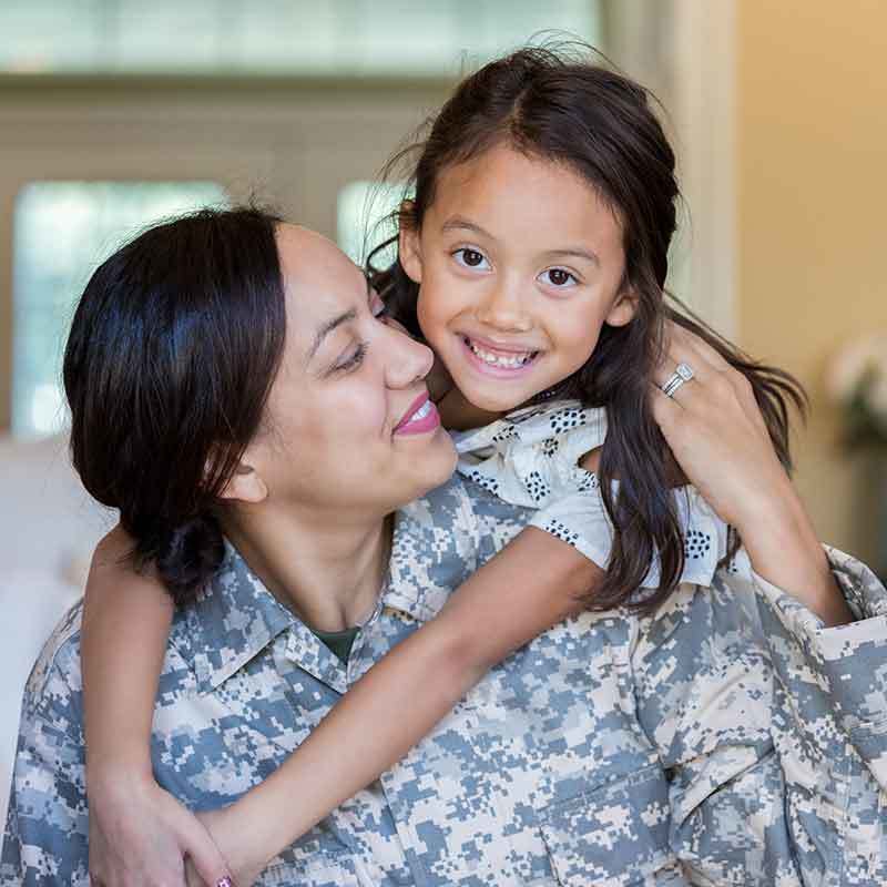 A mother wearing U.S. Army fatigues smiles at her daughter, draped over her mother's shoulder.