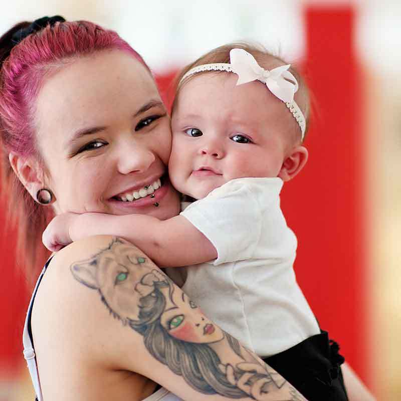 A smiling, young mother with pink hair and a tattoo holds her baby daughter.