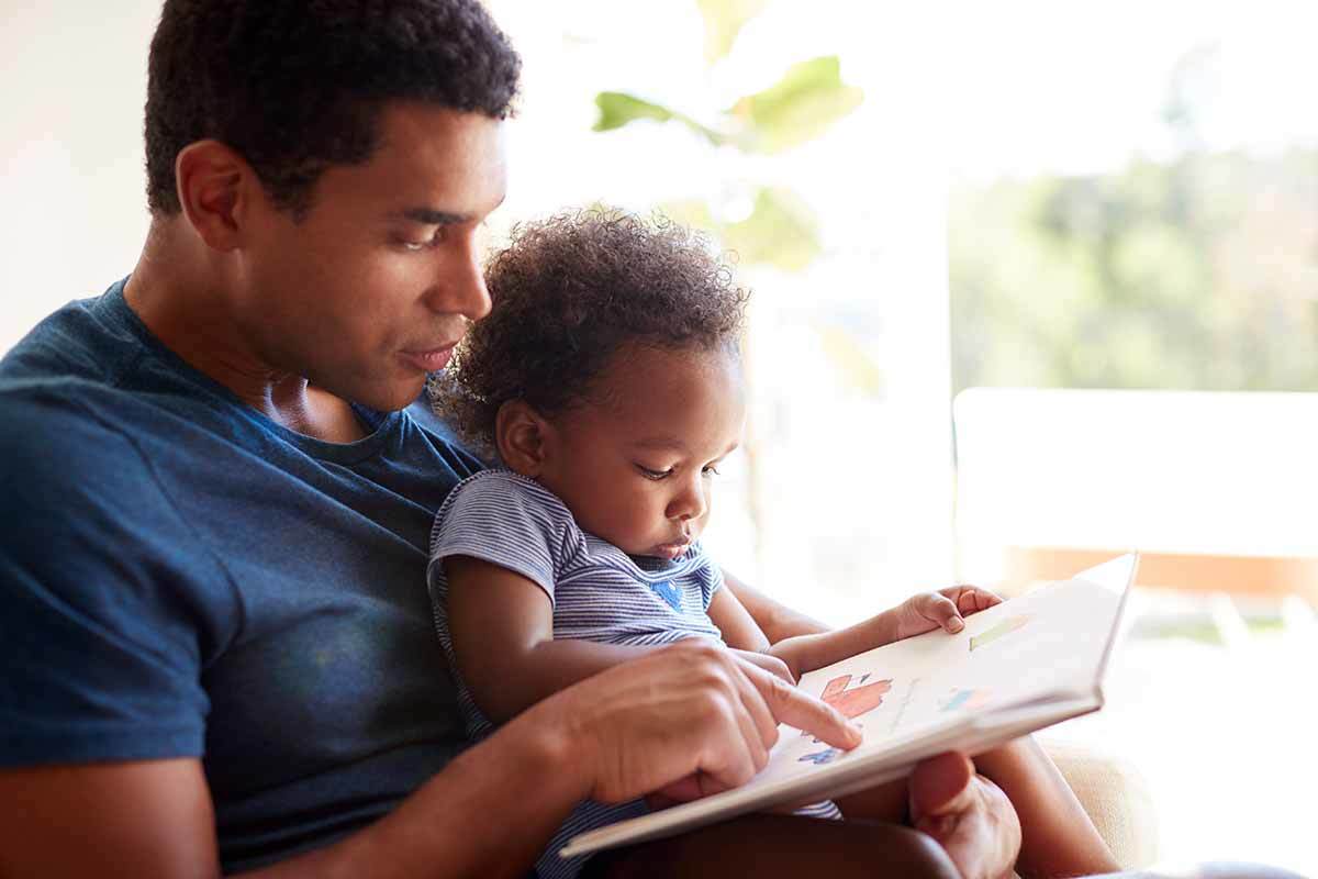 Close up of young adult black father reading a book with his two year old son, close up.